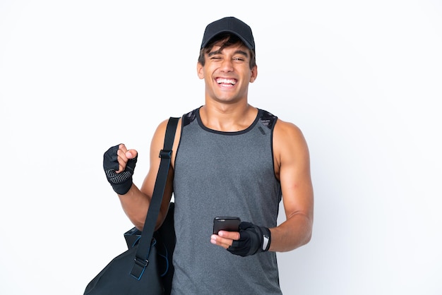 Deporte joven hombre caucásico con bolsa de deporte aislado sobre fondo blanco con teléfono en posición de victoria