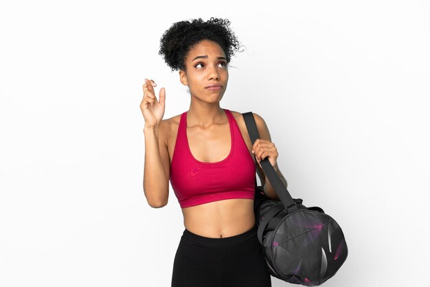 Deporte joven mujer afroamericana con bolsa de deporte aislado sobre fondo azul con los dedos cruzando y deseando lo mejor