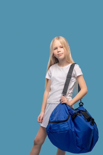 Deportes, entrenamiento. Linda chica de escuela primaria en camiseta blanca y falda de pie en una gran bolsa azul de deportes en su hombro