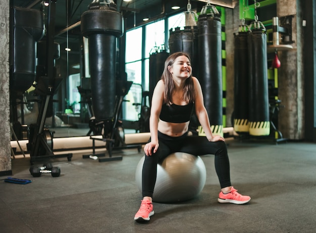 Deportes smilling mujer en ropa deportiva sentado en un fitball en un gimnasio oscuro.