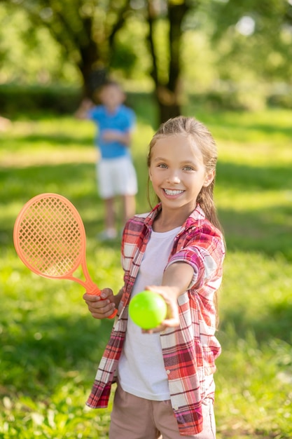 Deportes de verano. Sonriente chica rubia en camisa a cuadros con raqueta de tenis estirando la bola hacia adelante en el césped verde y los niños en la distancia