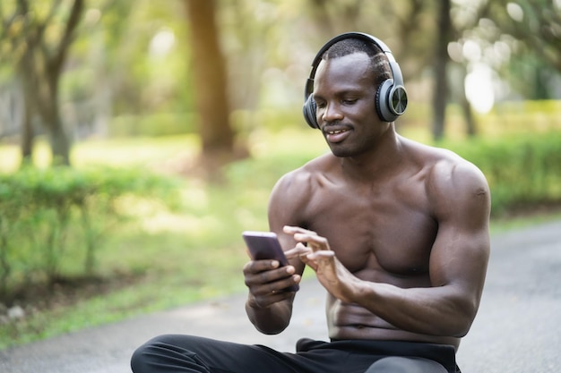 Deportista africano en forma de cuerpo mediante teléfono móvil y auriculares mientras descansa en el parque