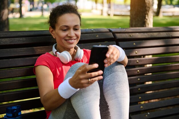 Deportista afroamericana en ropa deportiva con auriculares sonríe con dientes usando un teléfono inteligente y revisando la frecuencia cardíaca en la aplicación móvil mientras descansa después de hacer ejercicio al aire libre