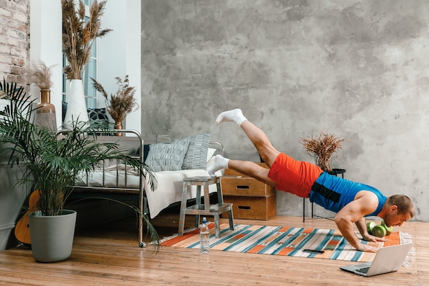 Deportista alegre con cabello negro hace flexiones y viendo ejercicios en línea desde la computadora portátil en el dormitorio