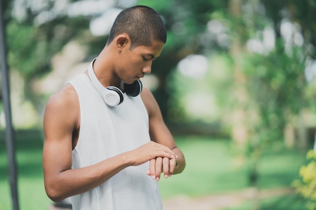 Deportista asiático en forma de cuerpo y pantalla táctil para reloj inteligente y usando auriculares blancos mientras descansa o antes de trotar haciendo ejercicio en el parque Concepto de carrera deportiva saludable