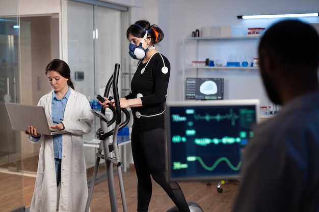 Deportista atleta con electrodos conectados a su cuerpo corriendo en la elíptica haciendo ejercicio cardiovascular. Investigador especialista sosteniendo un portátil analizando la resistencia controlando los datos de EGK en el laboratorio moderno