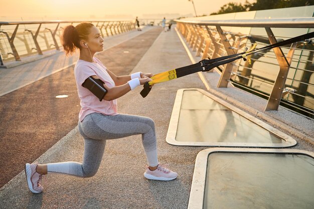 La deportista atlética hace ejercicios con correas de suspensión durante el entrenamiento de peso corporal en el entorno urbano de un puente de la ciudad. Atleta femenina de cuerpo completo en ropa deportiva tirando de cuerdas de suspensión y arremetiendo