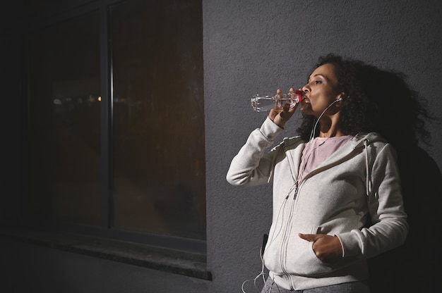 Deportista con auriculares en traje deportivo bebe agua apoyado en la pared gris