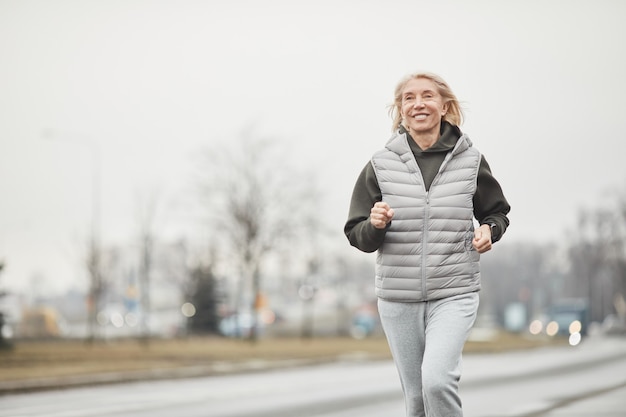 Deportista caucásica senior enérgica positiva en chaleco cálido y pantalón gris trotar a lo largo de la carretera mientras entrena en invierno