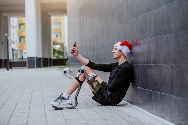 Deportista caucásico alegre con pierna artificial y con gorro de Papá Noel en la cabeza sentado en el suelo, apoyado en la pared y tomando selfie.