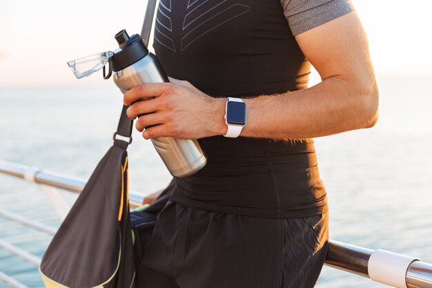 Deportista en chándal con bolsa deportiva caminando por el muelle o el paseo marítimo en la playa, en la mañana y sosteniendo la taza termo de metal