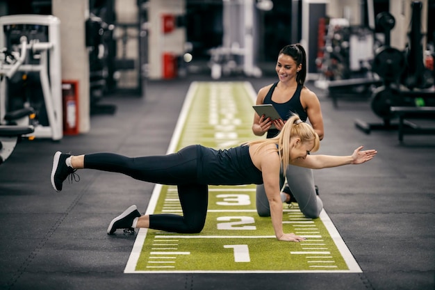 Una deportista está haciendo ejercicios en un gimnasio mientras su entrenadora personal está arrodillada junto a ella