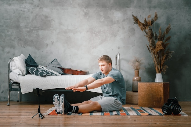 Foto el deportista estirándose, disparando blog, mira una película y las redes sociales en el dormitorio, al fondo hay una cama, un florero, una alfombra.