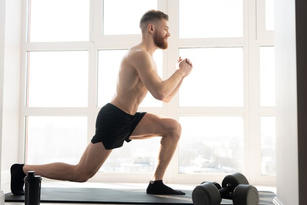 Deportista europeo haciendo estocadas ejercicio en colchoneta de fitness. Hombre barbudo joven con el torso desnudo. Concepto de actividad deportiva en casa. Interior del amplio apartamento moderno. Día soleado