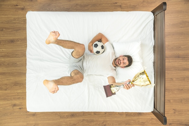 El deportista feliz yacía con una taza y una pelota en la cama. vista desde arriba