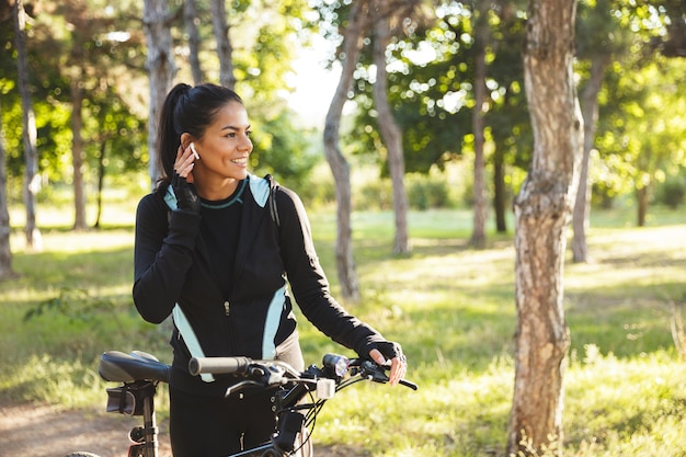 Deportista en forma atractiva con una bicicleta en el parque, escuchando música con auriculares inalámbricos