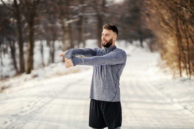 Deportista en forma haciendo ejercicios de calentamiento mientras está de pie en la naturaleza en un día nevado de invierno. Fitness de invierno, estilo de vida saludable