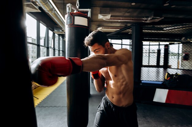 Deportista en guantes de boxeo rojos con un torso desnudo golpea el saco de boxeo en el gimnasio en el fondo del ring de boxeo.