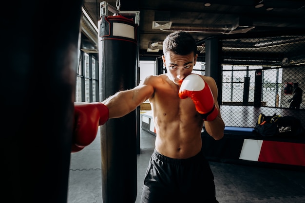 Deportista en guantes de boxeo rojos con un torso desnudo golpea el saco de boxeo en el gimnasio en el fondo del ring de boxeo.