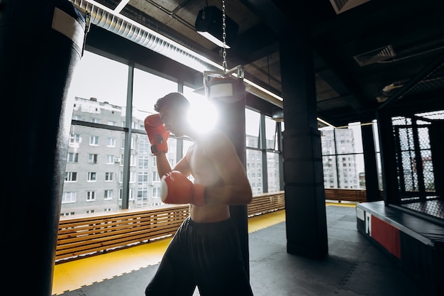 Deportista con guantes de boxeo rojos con un torso desnudo vestido con pantalones cortos negros golpea el saco de boxeo en el gimnasio con ventanas panorámicas.