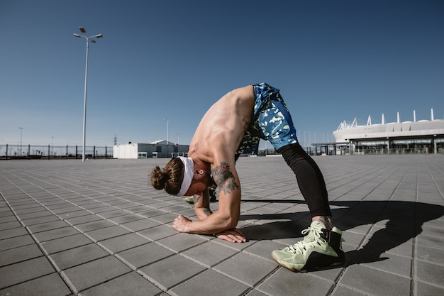 Deportista guapo sin camisa mirando a otro lado haciendo ejercicio de estiramiento en el campo de entrenamiento al aire libre en un día soleado. Estilo de vida deportivo.