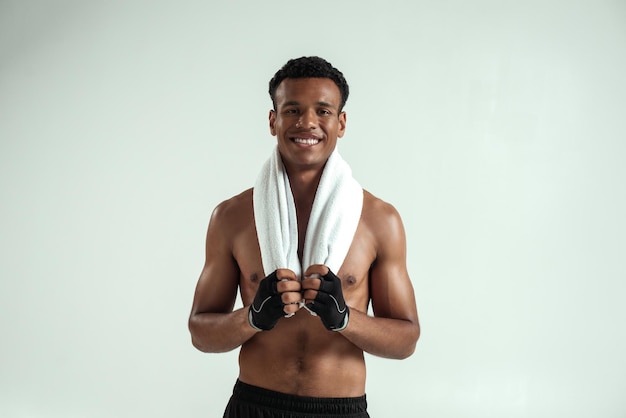 Deportista guapo. Joven africano con una toalla sobre los hombros y el torso desnudo mirando a la cámara y sonriendo mientras está de pie contra el fondo gris. Concepto de deporte. Tiro del estudio