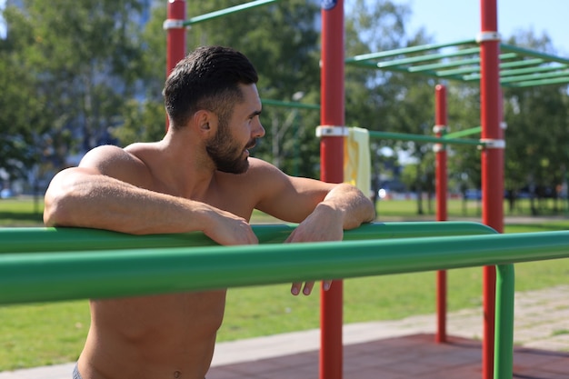 Deportista guapo con el torso desnudo descansando mientras hace ejercicio en un día soleado en el parque.