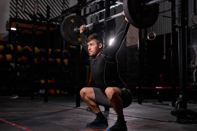 Deportista haciendo ejercicio con barra. Joven culturista masculino musculoso caucásico haciendo ejercicios de levantamiento de pesas en el gimnasio oscuro, utilizando equipo deportivo