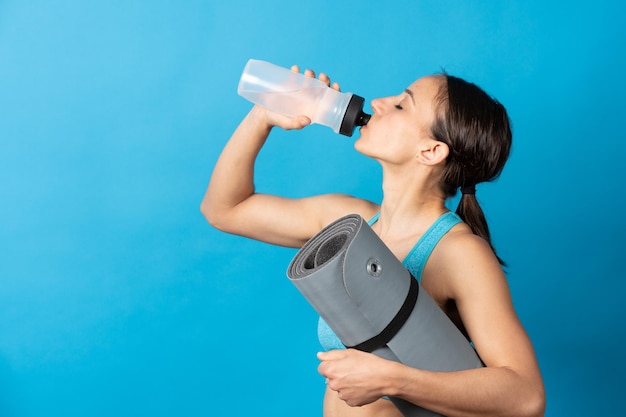 Deportista hispana bebiendo agua y sosteniendo estera