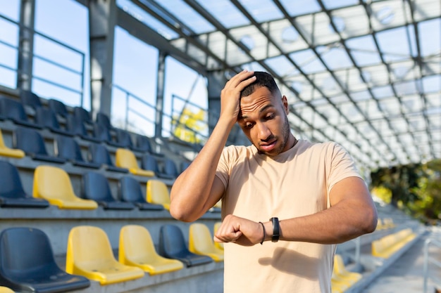 Deportista hispano molesto parado en el estadio sosteniendo la cabeza y mirando el reloj inteligente de pulsera