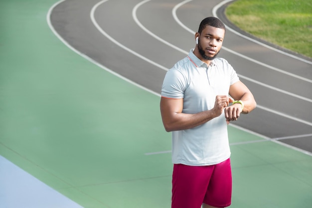 Deportista joven en forma seria con un rastreador de fitness de pie en la pista de atletismo en el estadio mientras mira hacia otro lado