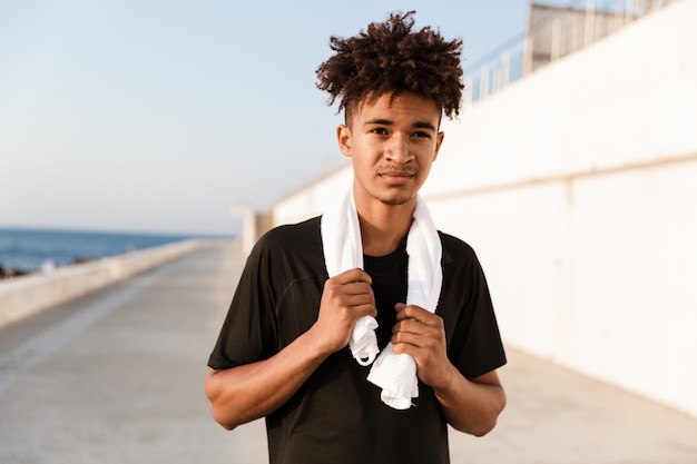 Deportista joven guapo de pie con una toalla al aire libre en la playa