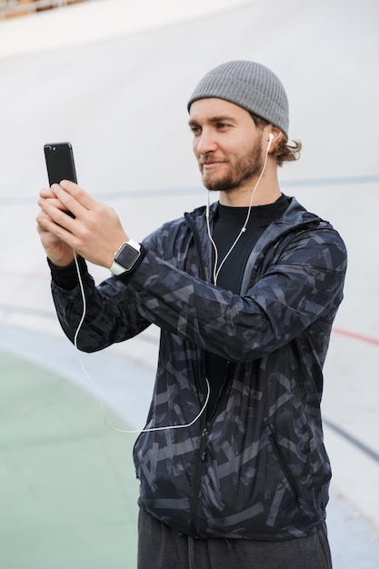 Deportista joven sonriente motivado escuchando música con auriculares mientras está de pie en el estadio, tomando un selfie con teléfono móvil