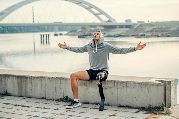 Un deportista libre con pierna protésica con sudadera con capucha sentado en el muelle