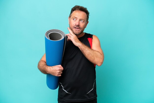 Deportista de mediana edad que va a clases de yoga mientras sostiene una alfombra aislada de fondo azul mirando hacia arriba mientras sonríe