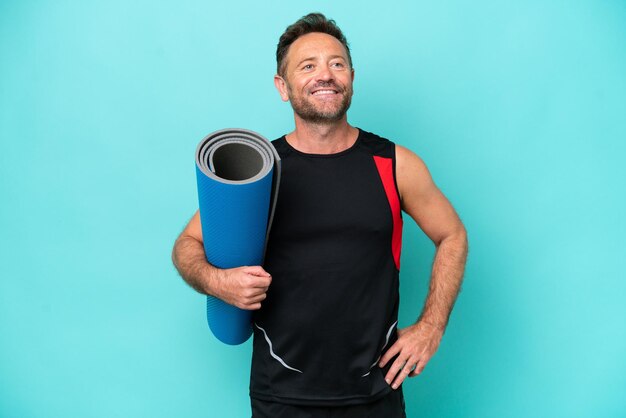 Deportista de mediana edad yendo a clases de yoga mientras sostiene una alfombra aislada de fondo azul posando con los brazos en la cadera y sonriendo