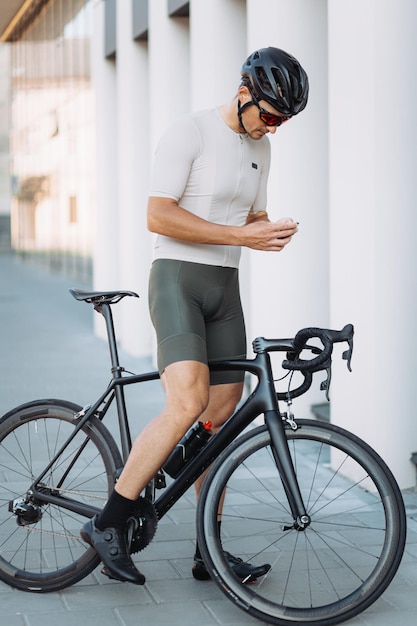 Deportista musculoso parado al aire libre con su bicicleta y sosteniendo un navegador multimedia en las manos Ciclista profesional usando un aparato moderno para entrenar en el área urbana