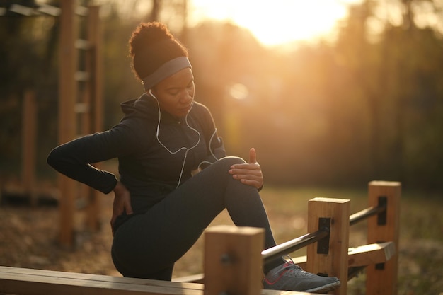 Deportista negra sintiendo dolor después de lesionarse la rodilla durante el entrenamiento deportivo en el parque al atardecer