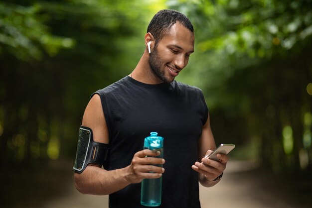 Foto deportista negro sonriente haciendo ejercicio en el parque bebiendo y usando un teléfono inteligente