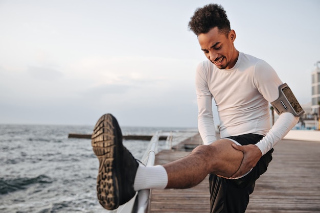 Deportista en pantalones cortos negros y camiseta blanca de manga larga hace auto masaje