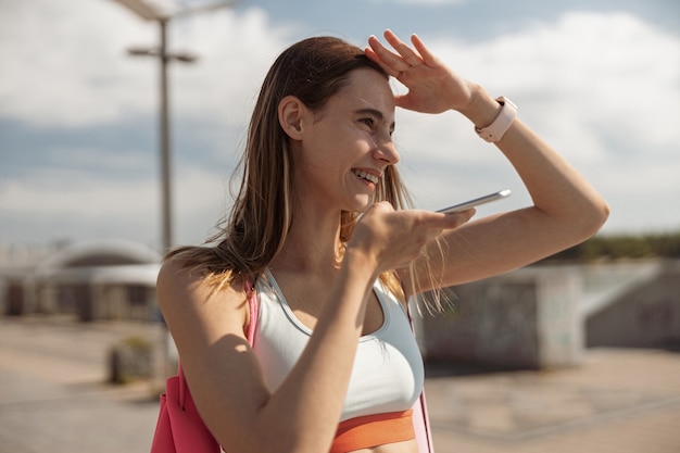 Deportista en la parte superior usa el modo de altavoz fuerte en el teléfono protegiéndose los ojos con la mano