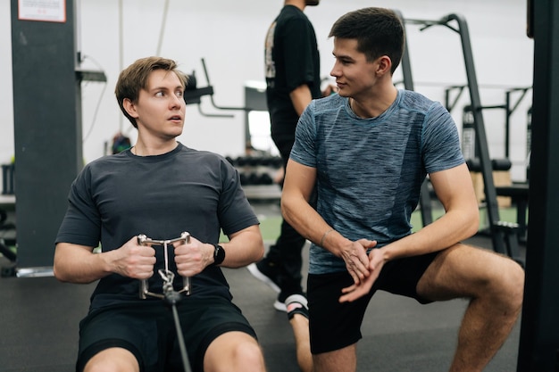 Deportista principiante con dificultades para practicar en una máquina de remo durante el entrenamiento deportivo con un entrenador personal en un club de salud Atleta masculino haciendo ejercicios con correa en una maquinaria de remo