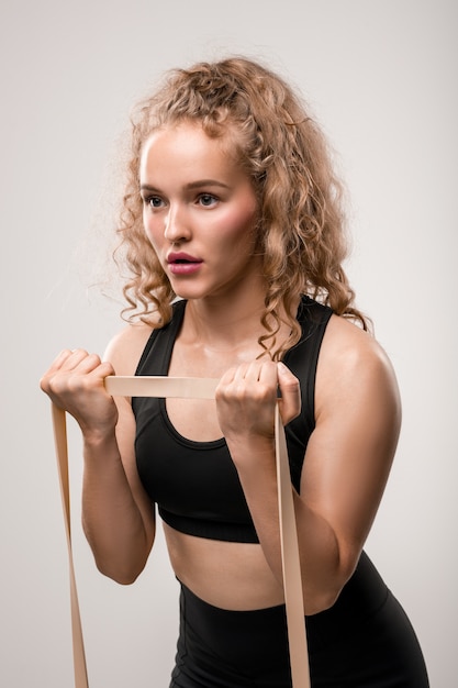 Deportista rubia rizada con banda flexible haciendo ejercicio físico para brazos durante el entrenamiento diario en el gimnasio o en casa