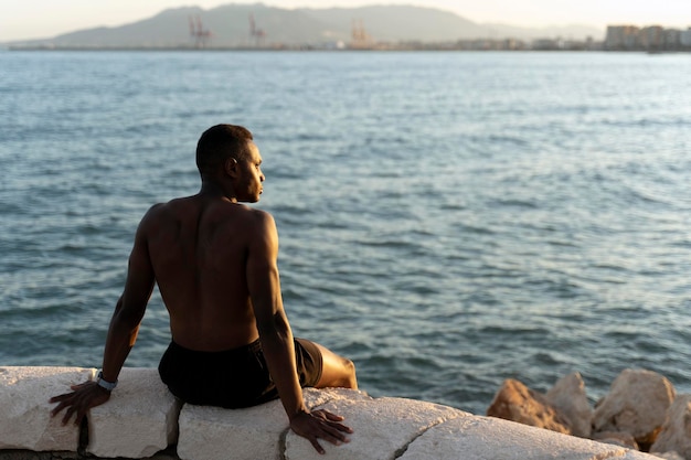 Deportista sentado en la costa del océano y mirando hacia otro lado mientras disfruta de la puesta de sol