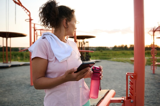 Deportista con smartphone y botella de agua en el campo deportivo al atardecer