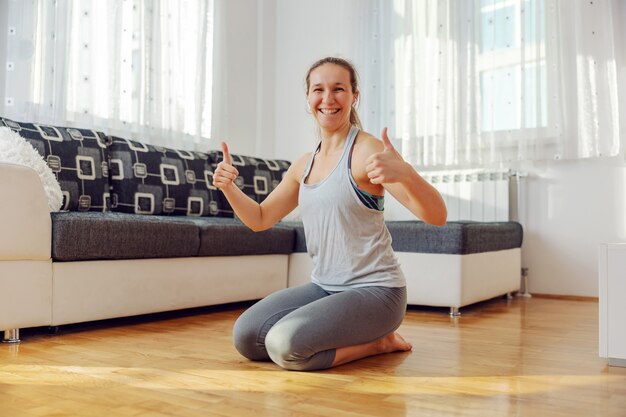 Deportista sonriente arrodillado en el suelo en casa y mostrando los pulgares hacia arriba.