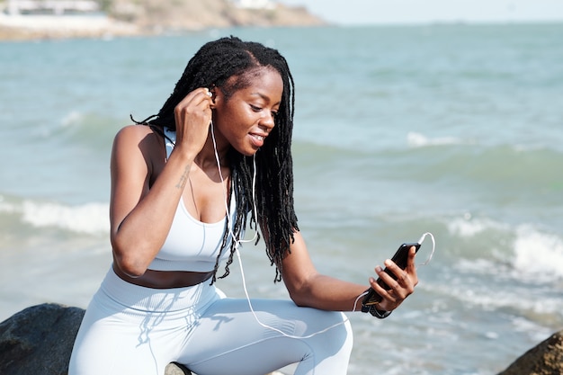 Deportista sonriente sentado en la playa rocosa y ponerse auriculares al hacer una videollamada a un amigo o entrenador