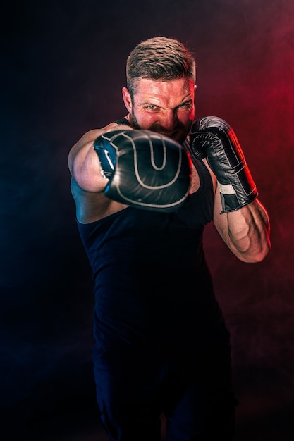 Deportista tatuado barbudo muay thai boxeador en camiseta negra y guantes de boxeo peleando sobre fondo oscuro con humo. Concepto de deporte.