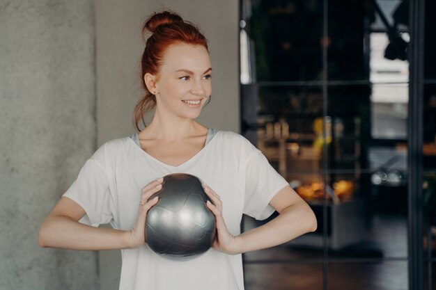 Deportiva buena mujer con el pelo rojo con ropa deportiva sosteniendo una pequeña bola con las dos manos y sonriendo a alguien, lista para entrenar en el gimnasio. Concepto de salud y cuidado personal.