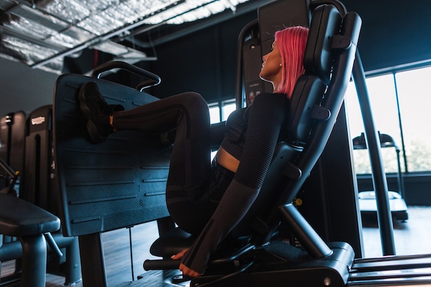 Deportiva hermosa joven con cabello rosado en ropa deportiva negra con zapatillas de deporte se sienta en el simulador y bombea sus piernas